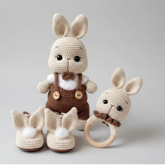 Set of crocheted baby items including a bunny outfit, shoes, and teether on a textured surface.