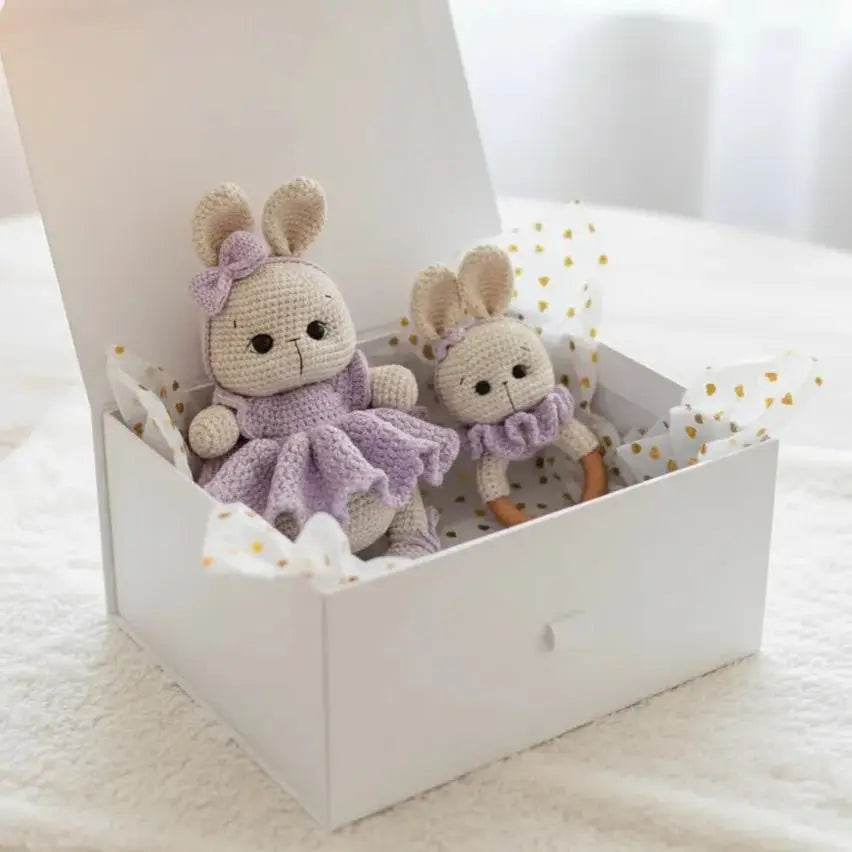 Two crocheted bunny dolls in lavender and beige, sitting in a white gift box with gold-dotted tissue paper.
