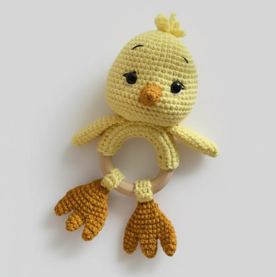 Crocheted yellow chick baby rattle with wooden ring, sitting among green leaves