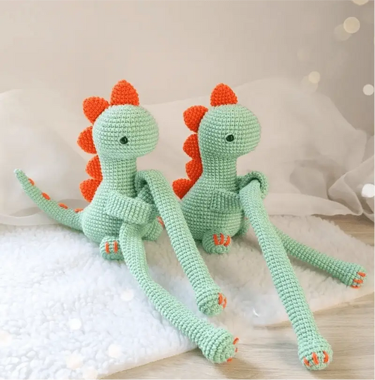 Two mint green crocheted dinosaurs with orange spikes sitting on a white surface.
