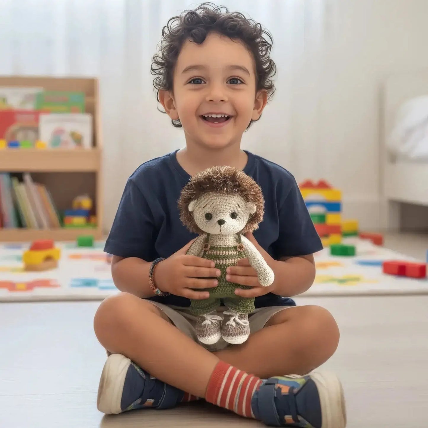 Smiling child sits cross-legged, holding crocheted hedgehog toy in playroom with blocks