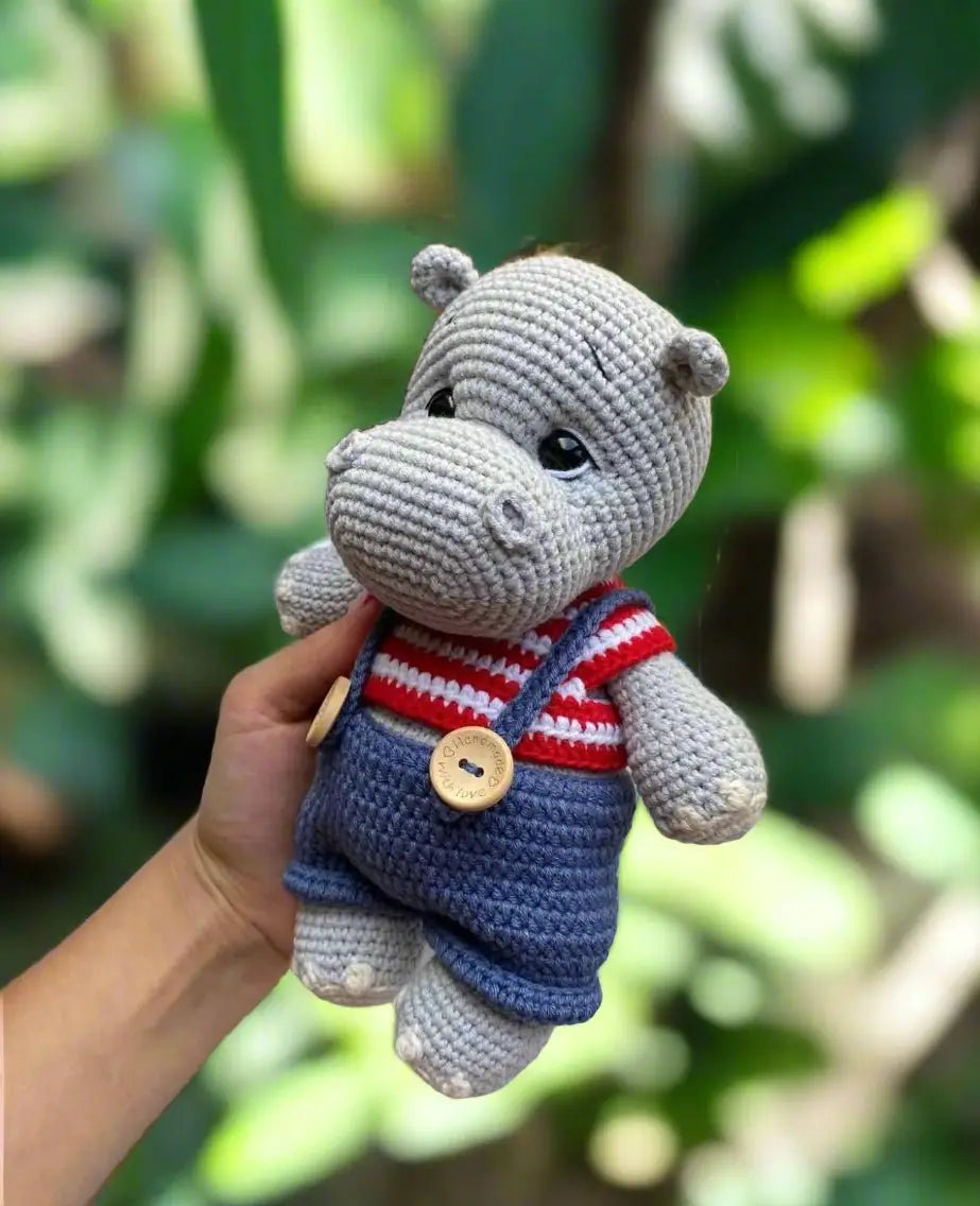 Crocheted gray hippo toy in blue overalls and red-striped shirt, held by a hand