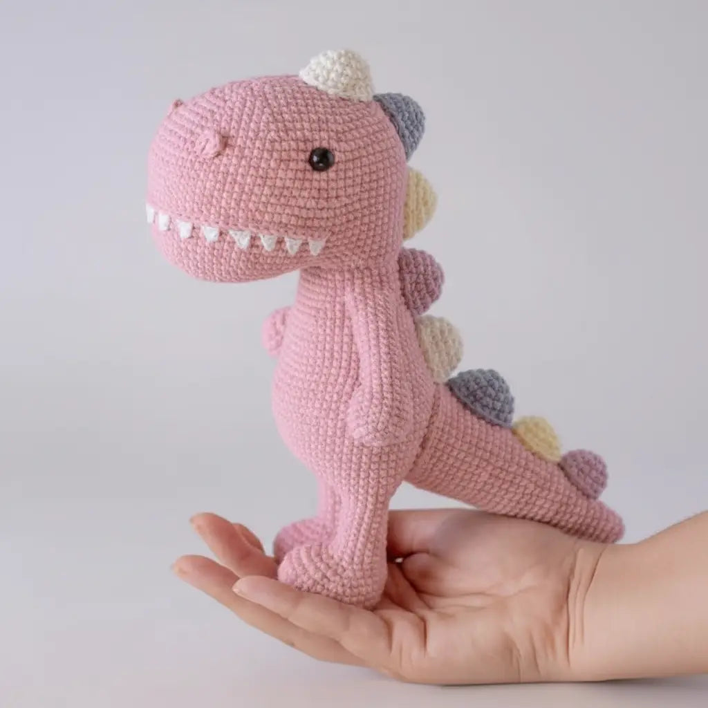 Pink crocheted dinosaur with sharp teeth sitting on an open palm, multicolored spiky back