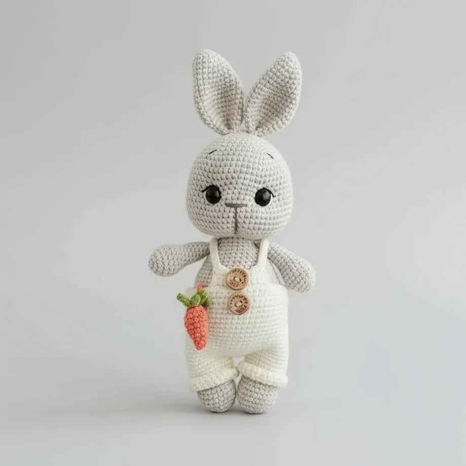 Crocheted bunny in white overalls holding a carrot, standing against a gray background.