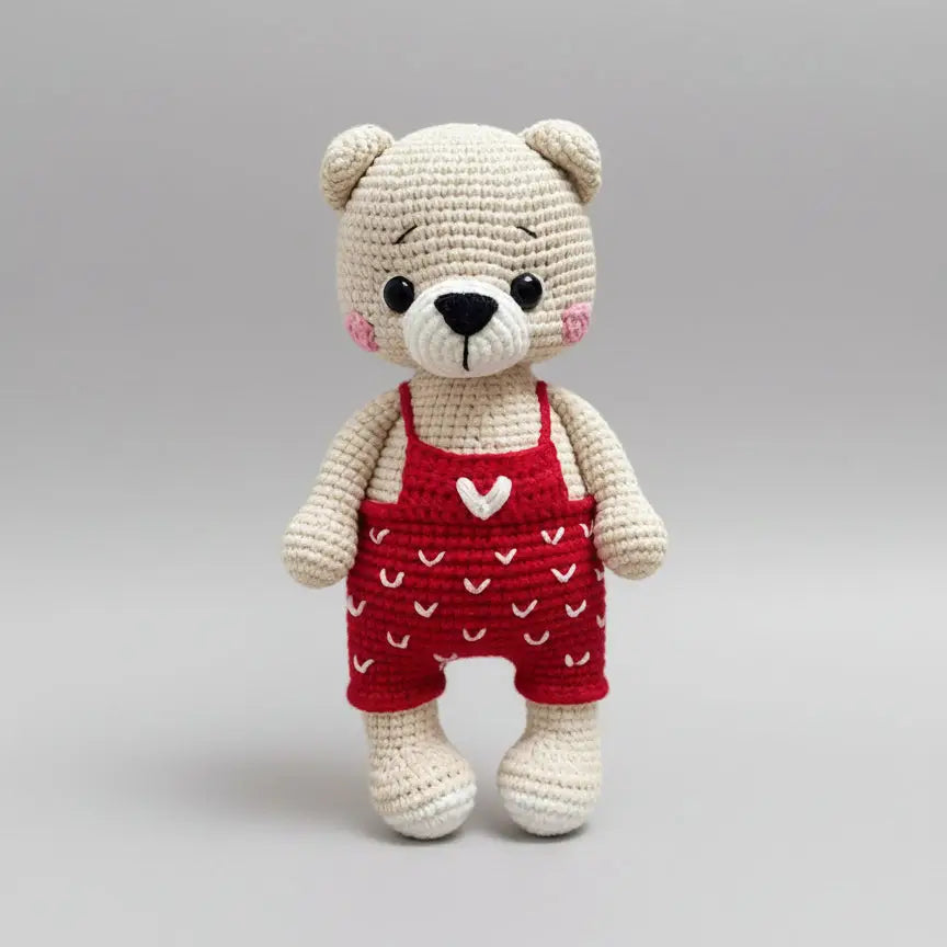 Crocheted teddy bear in red overalls with white heart details, standing cute