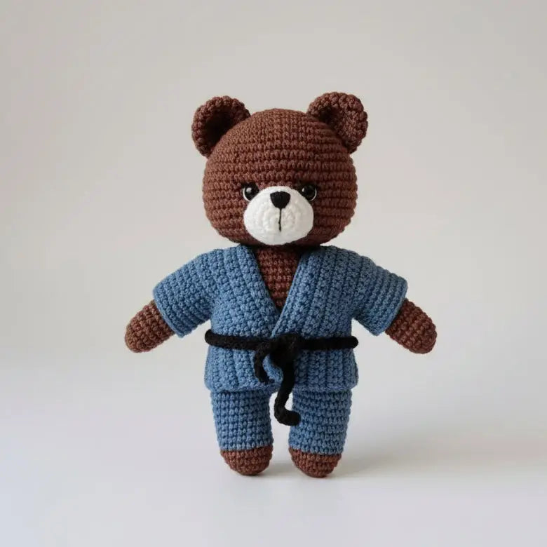 Crocheted brown teddy bear wearing blue bathrobe and black belt, standing upright