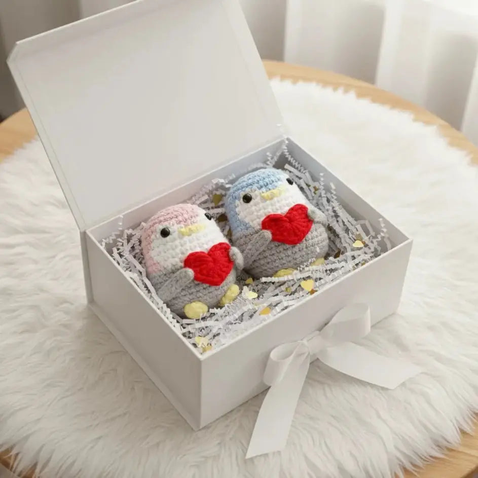 Two crocheted penguin dolls with red hearts in a white gift box with a ribbon