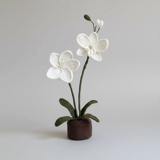Crocheted white orchid with green leaves in a dark brown pot against a neutral background