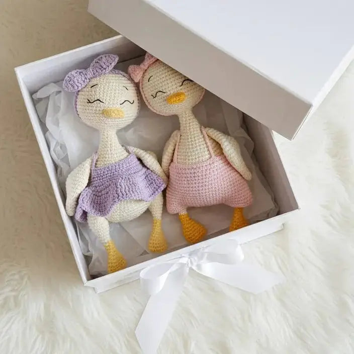 Two crocheted duck dolls in purple and pink outfits nestled in a white gift box with tissue paper