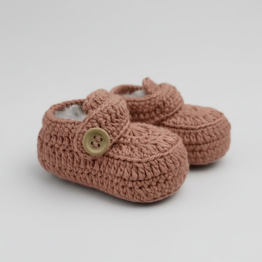 Dusty pink crocheted baby booties with wooden button, resting on blurred green background