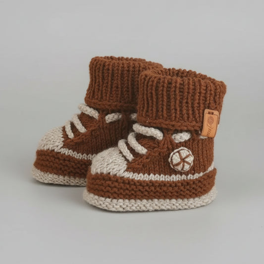 Handknit brown and cream baby booties with laces and side logo, nestled on soft fabric