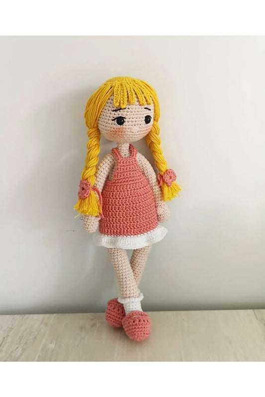 Crocheted doll with blonde braids, pink dress, and white trim, sitting against a white wall