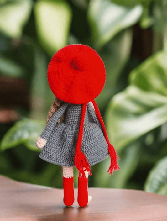 Crocheted doll in red beret and gray coat, standing against blurry green leaves