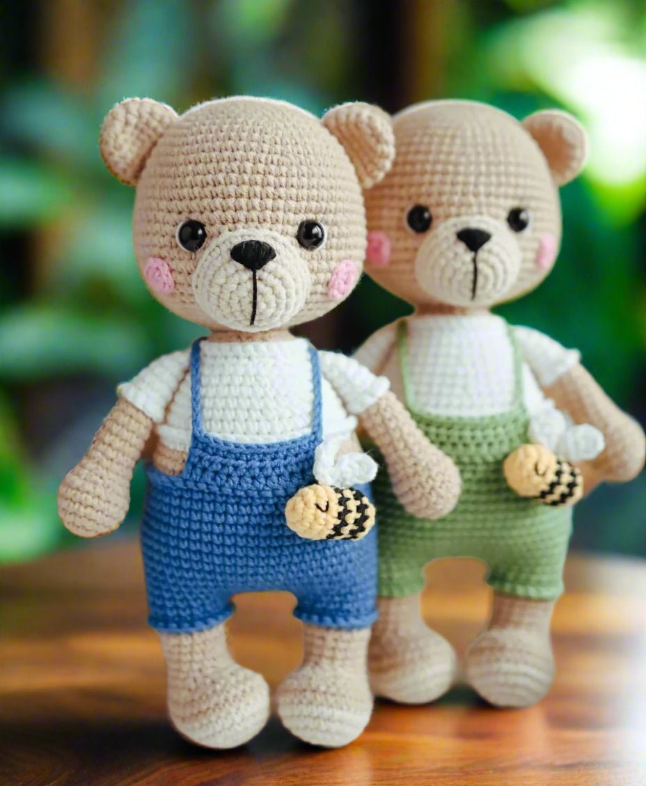Two crocheted teddy bears in blue and green overalls holding cookies, with rosy cheeks