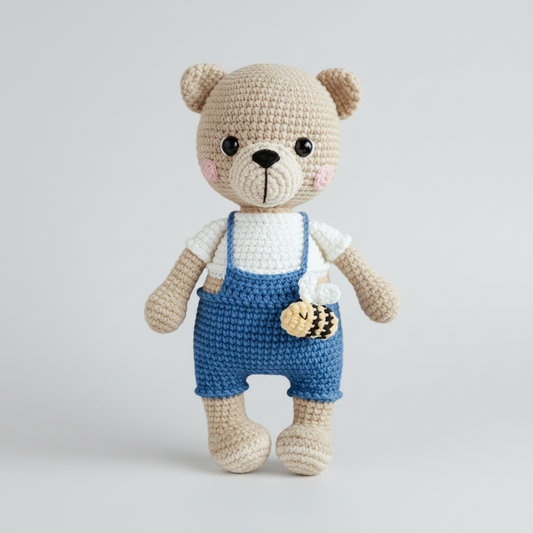 Crocheted teddy bear in blue overalls, holding a bee, against a light gray background