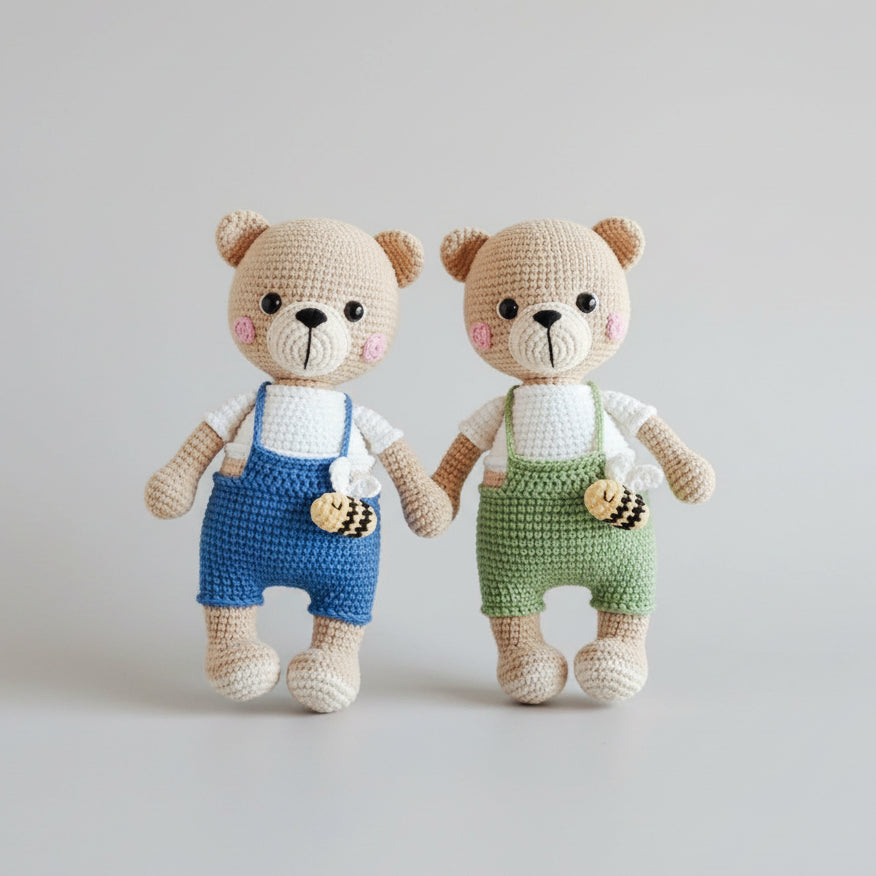 Two crocheted teddy bears wearing blue and green overalls, surrounded by green leaves.