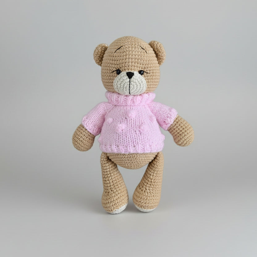 Crocheted teddy bear wearing a pink knitted sweater, standing against a light background