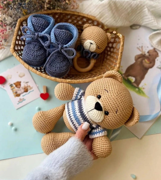 Crocheted baby booties and teddy bear in a woven basket, with a hand holding the bear