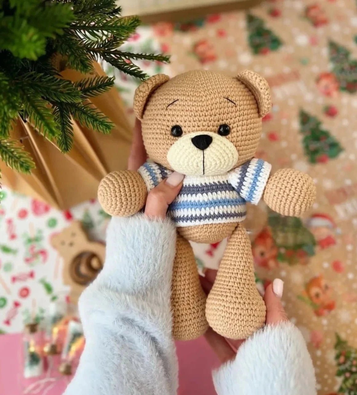 Crocheted teddy bear in blue-white striped sweater, held near a Christmas tree branch