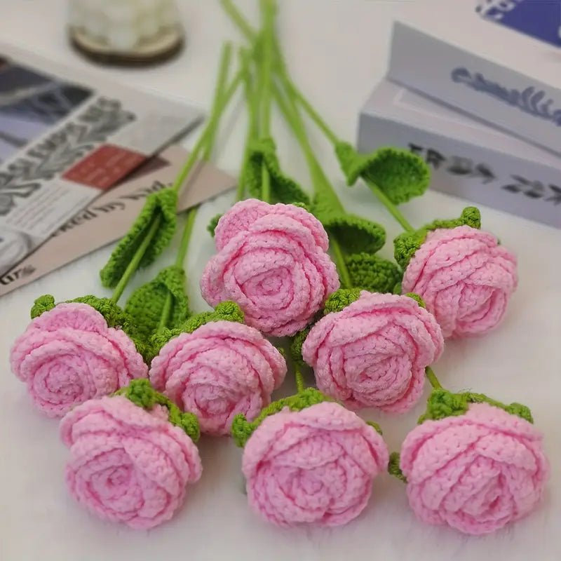 Crocheted pink roses with green leaves on a white surface with books and newspapers