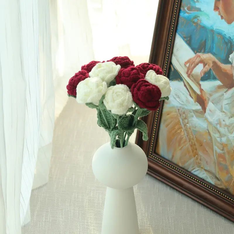 White vase with red and white crocheted roses near framed artwork and lace curtain