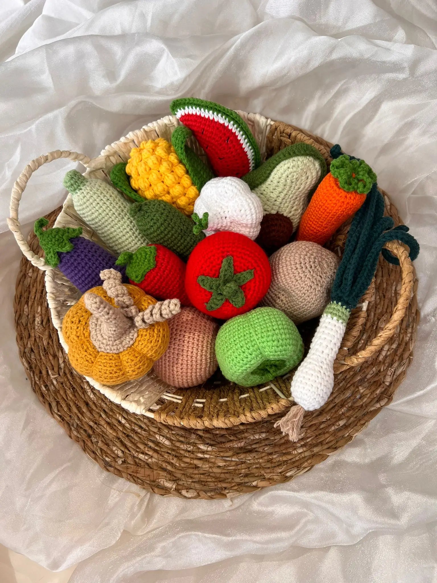 Crocheted fruits and vegetables in a woven basket on a white fabric background.