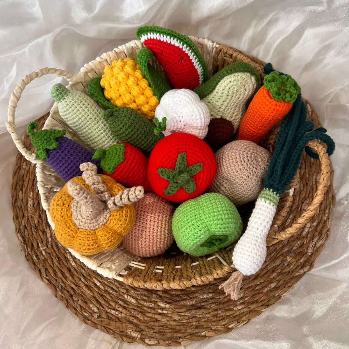 Crocheted fruits and vegetables in a woven basket on a white fabric background.