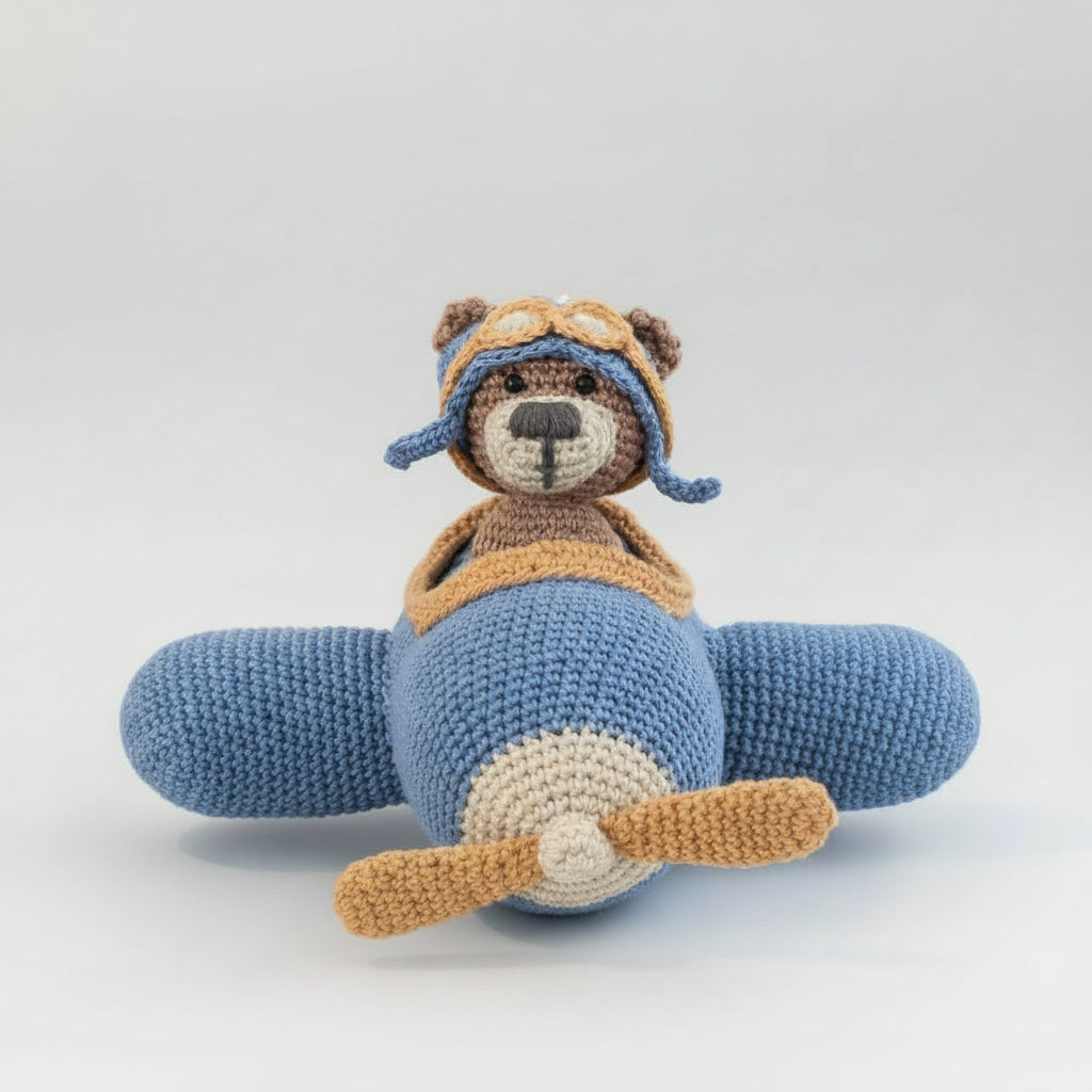 A crocheted teddy bear wearing a blue and tan aviator hat and sitting in a blue toy airplane on a brown fabric couch. The airplane has a wooden propeller and a small wooden bead on a string.