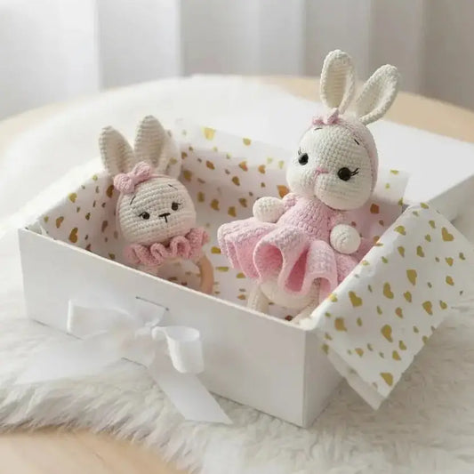 Two crocheted bunny dolls in pink tutus sit inside a heart-patterned gift box on fluffy fabric.