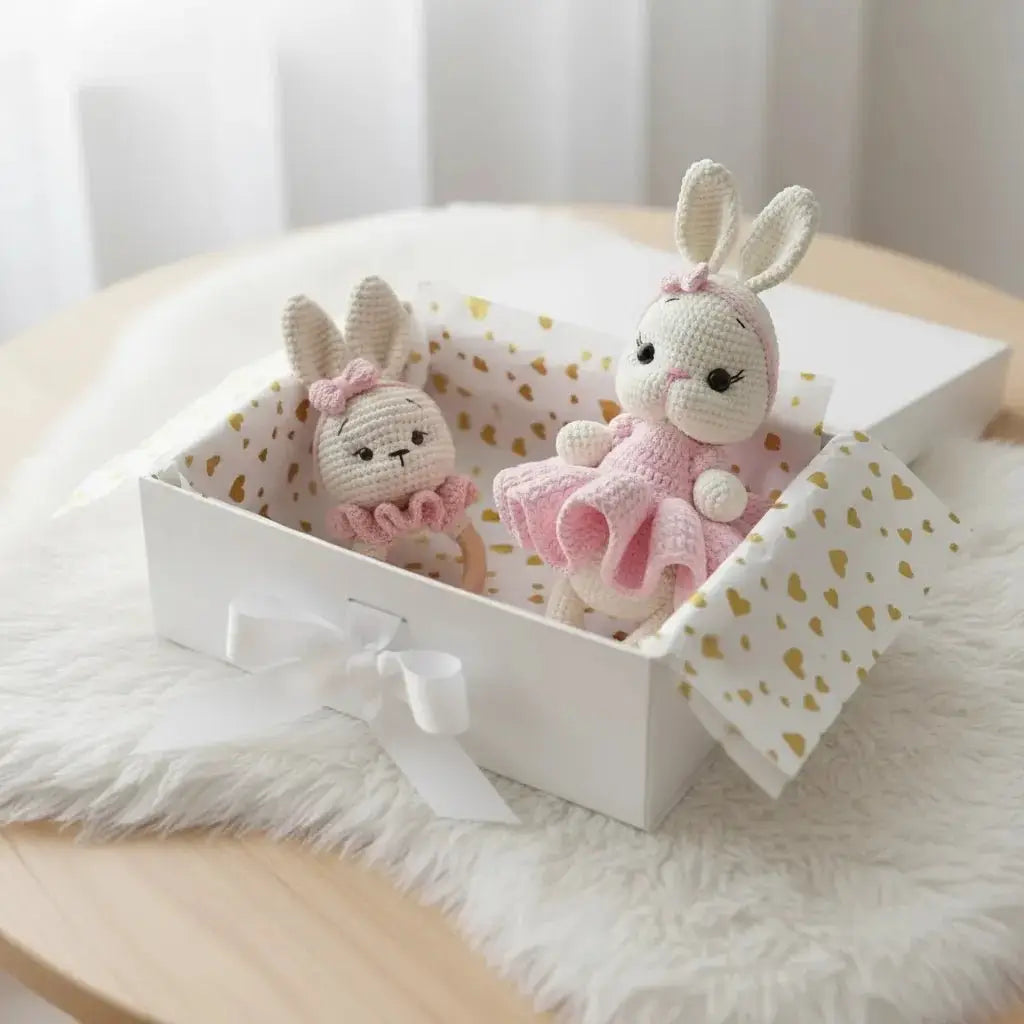 Two crocheted bunny dolls in pink tutus sit inside a heart-patterned gift box on fluffy fabric.