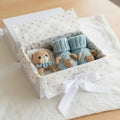 Knitted blue baby booties and teddy bear in a white gift box with gold hearts