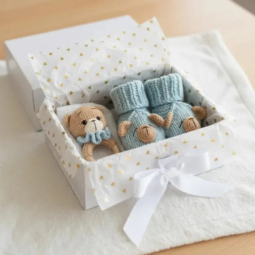 Knitted blue baby booties and teddy bear in a white gift box with gold hearts