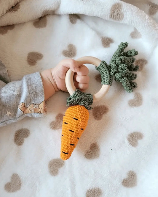 Crocheted carrot teething ring held by baby's hand on spotty blanket