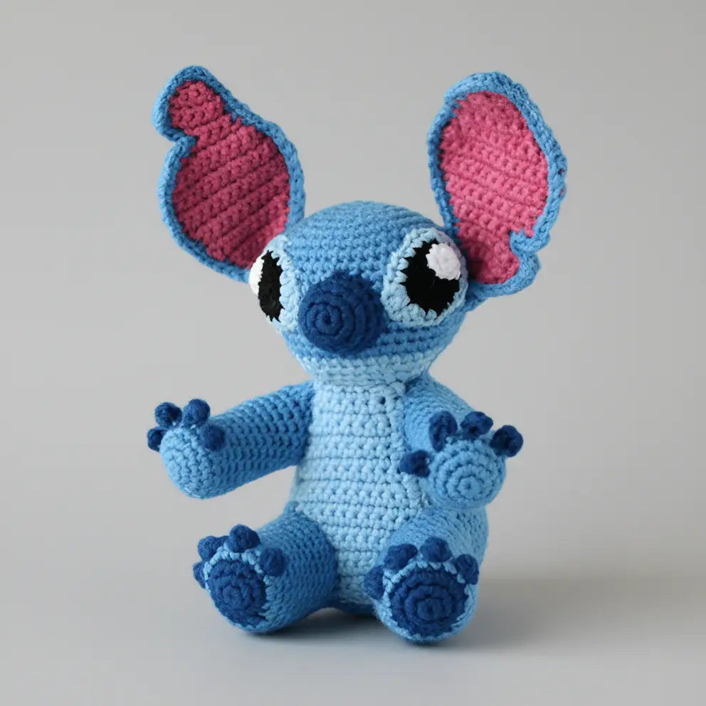 Blue crocheted creature with large pink-lined ears and spiral-patterned paws, sitting upright.