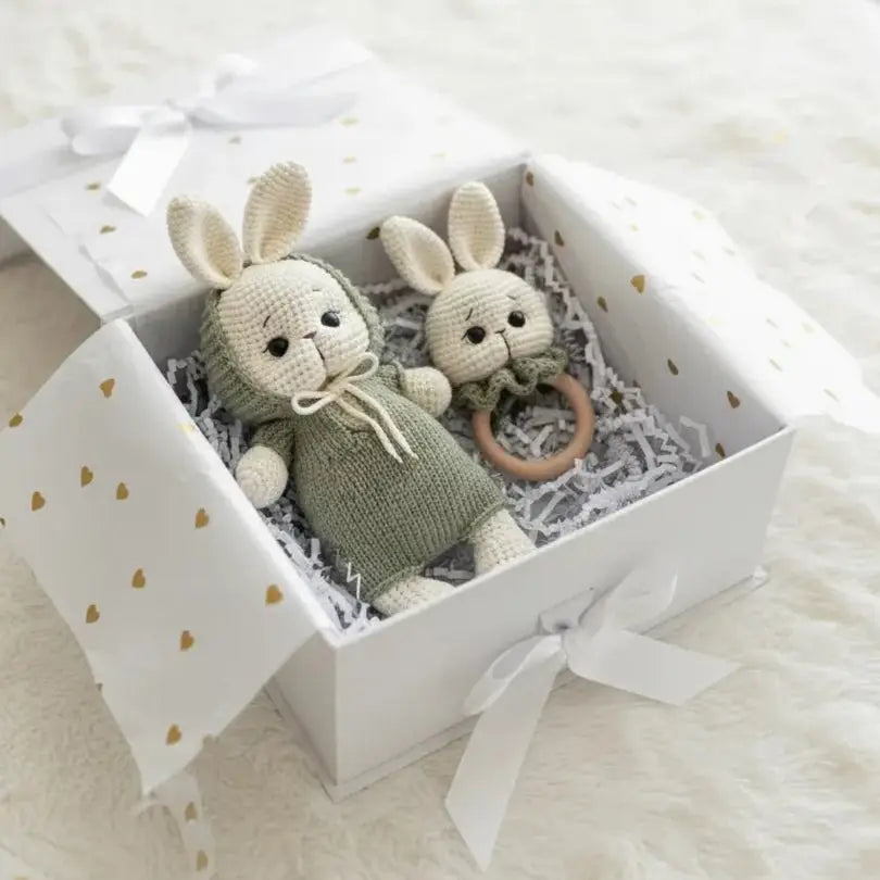 Two knitted bunnies in a gift box with gold heart pattern, nestled in shredded paper