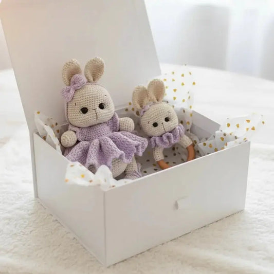Two crocheted bunny dolls in lavender and beige, sitting in a white gift box with gold-dotted tissue paper.