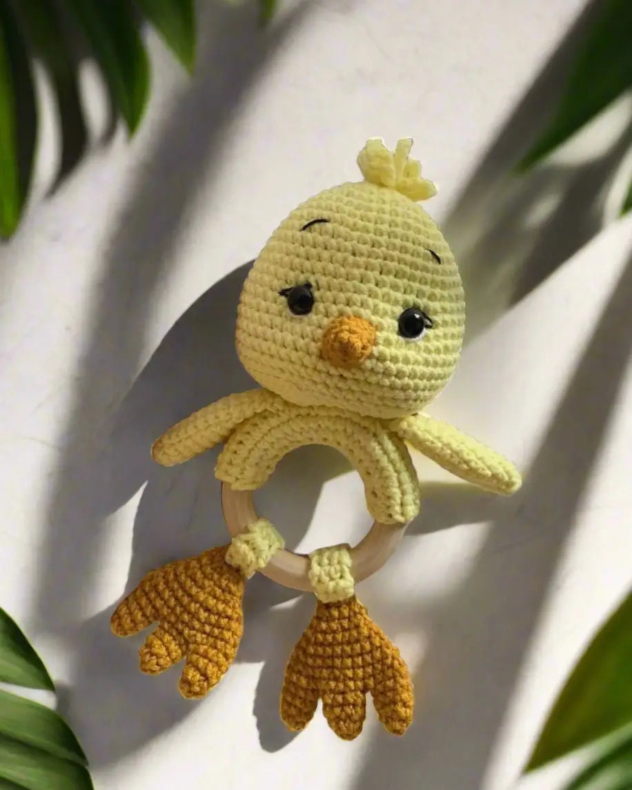 Crocheted yellow chick baby rattle with wooden ring, sitting among green leaves