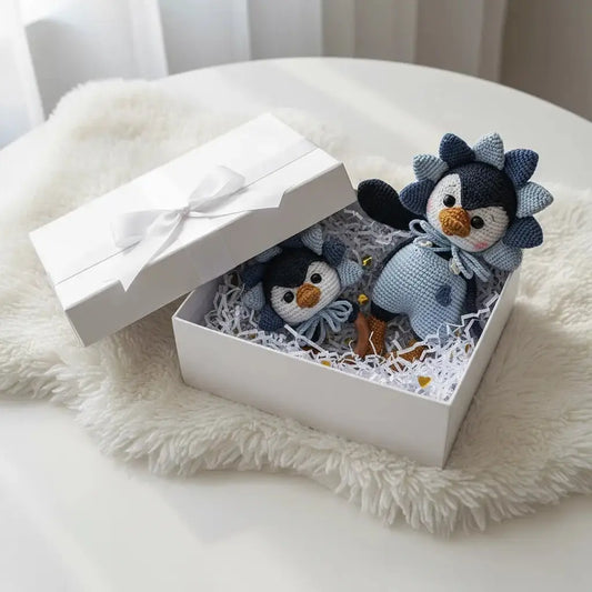 Two crocheted penguin toys nestled in a white gift box with shredded paper, sitting on a fluffy white surface.