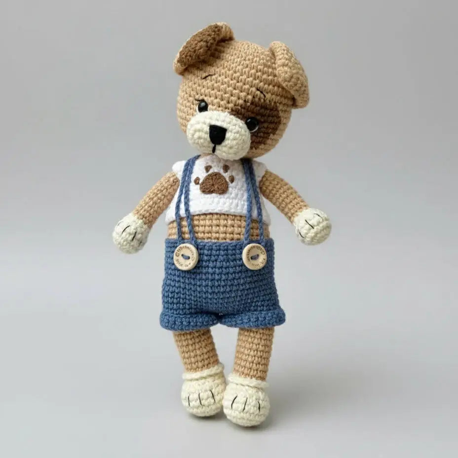 Crocheted teddy bear in blue overalls with paw print, standing on gray background