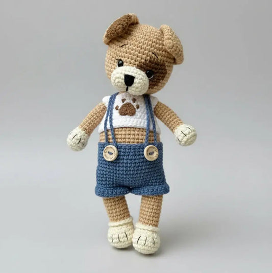 Crocheted teddy bear in blue overalls with paw print, standing on gray background
