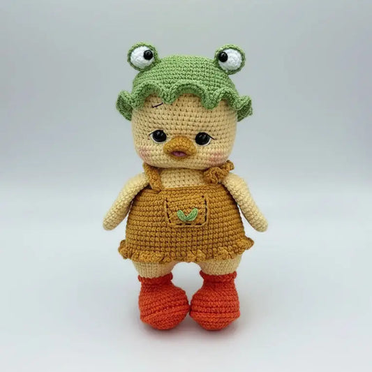 Crocheted chick doll with frog hat, yellow and orange, standing on neutral background