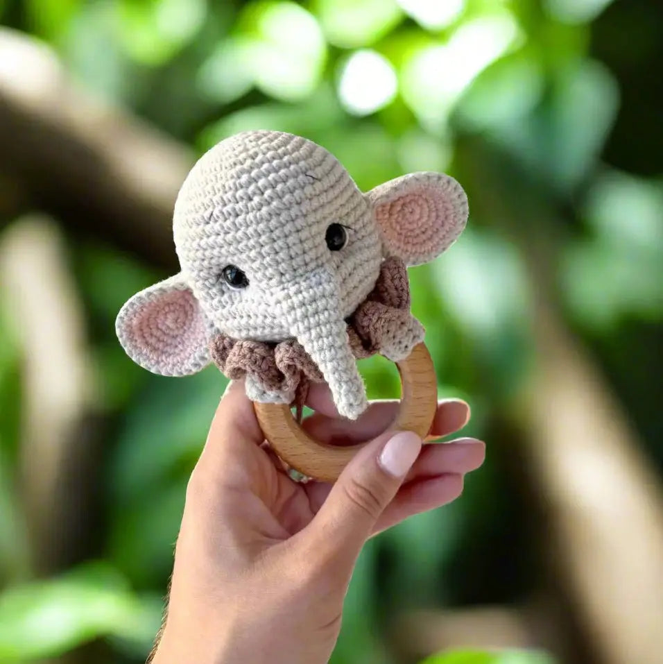 Crocheted elephant baby rattle with wooden ring, held against green blurred background