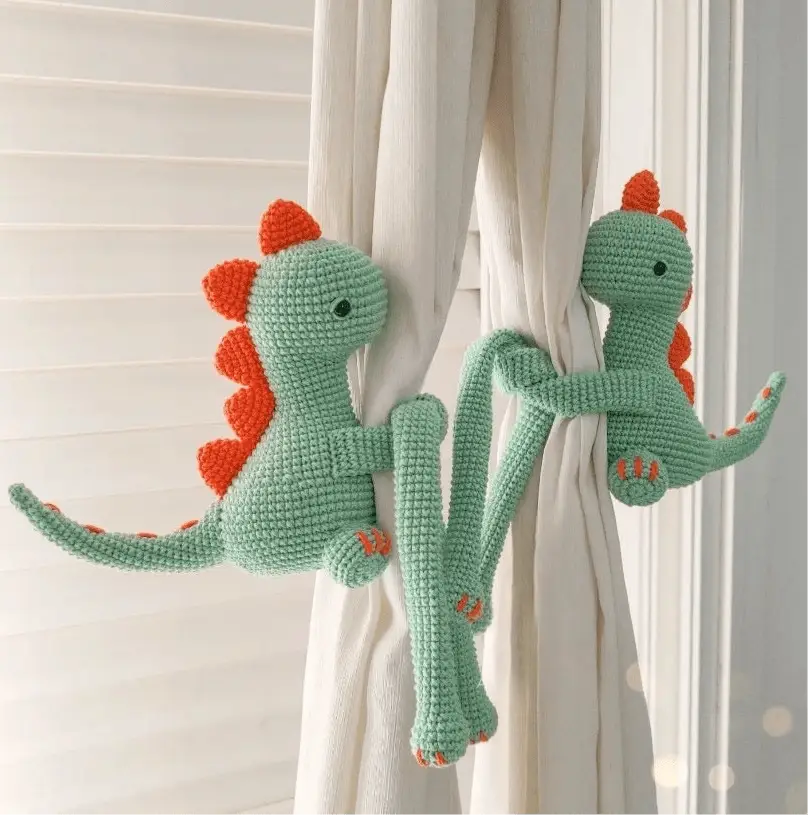 Two crocheted green and orange dinosaurs hanging on white curtains, playfully posed together.