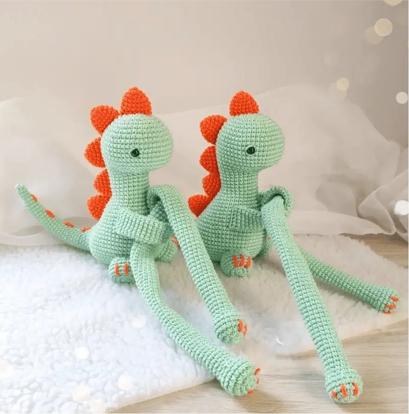 Two mint green crocheted dinosaurs with orange spikes sitting on a white surface.