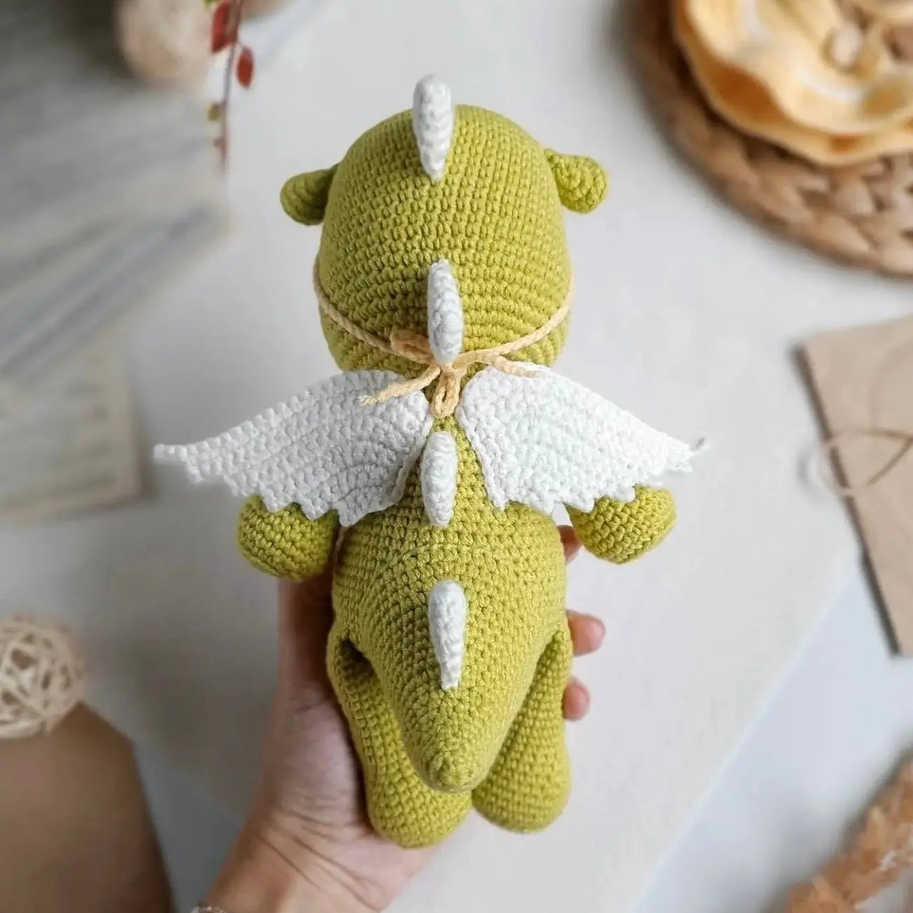 Green crocheted dragon with white wings, held in a hand with a bracelet.