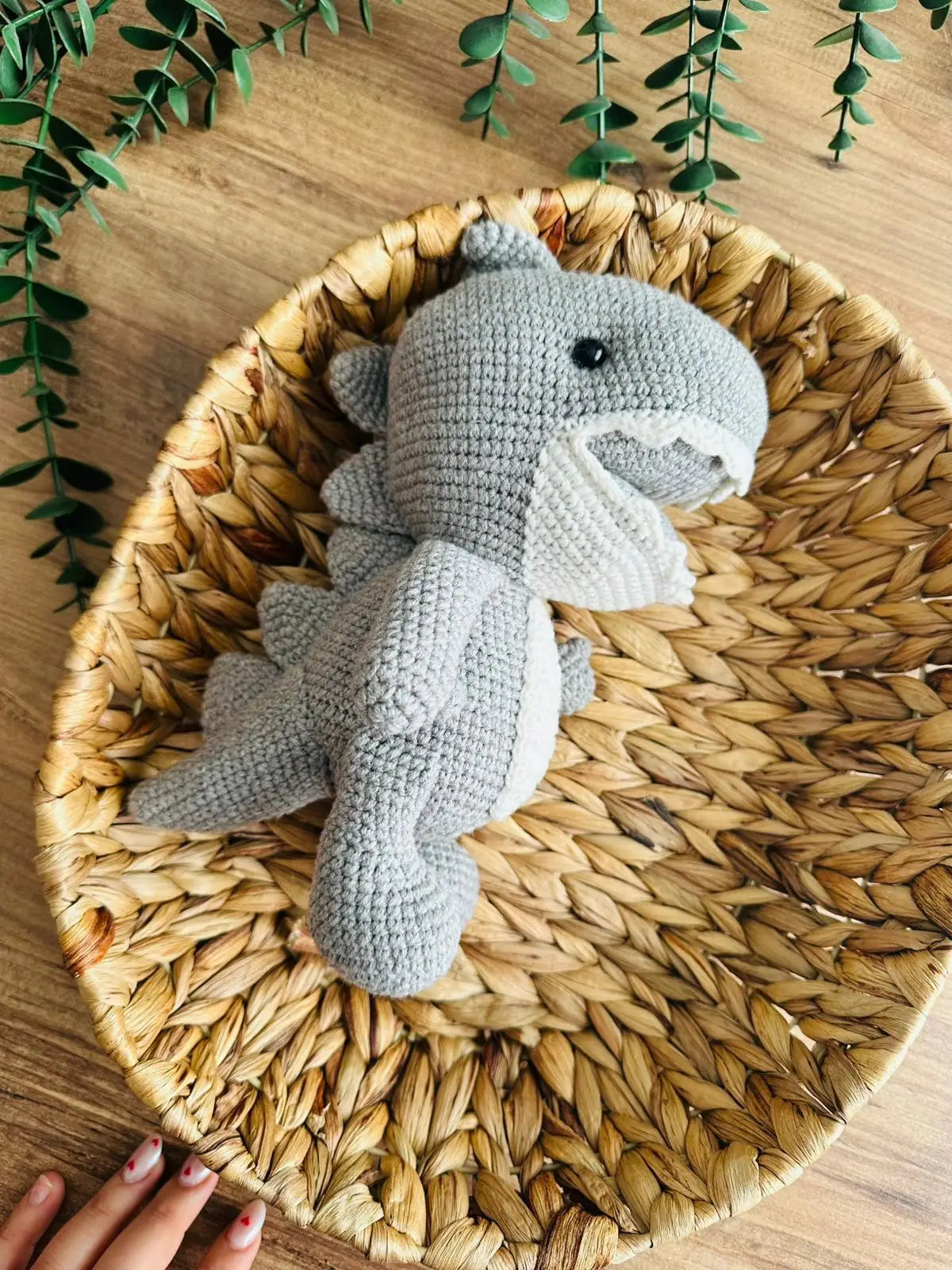 Gray crocheted shark plush toy sitting in a woven basket with green plant leaves