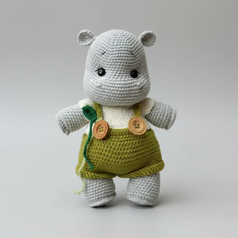 Crocheted gray hippo wearing green overalls with wooden buttons on neutral background