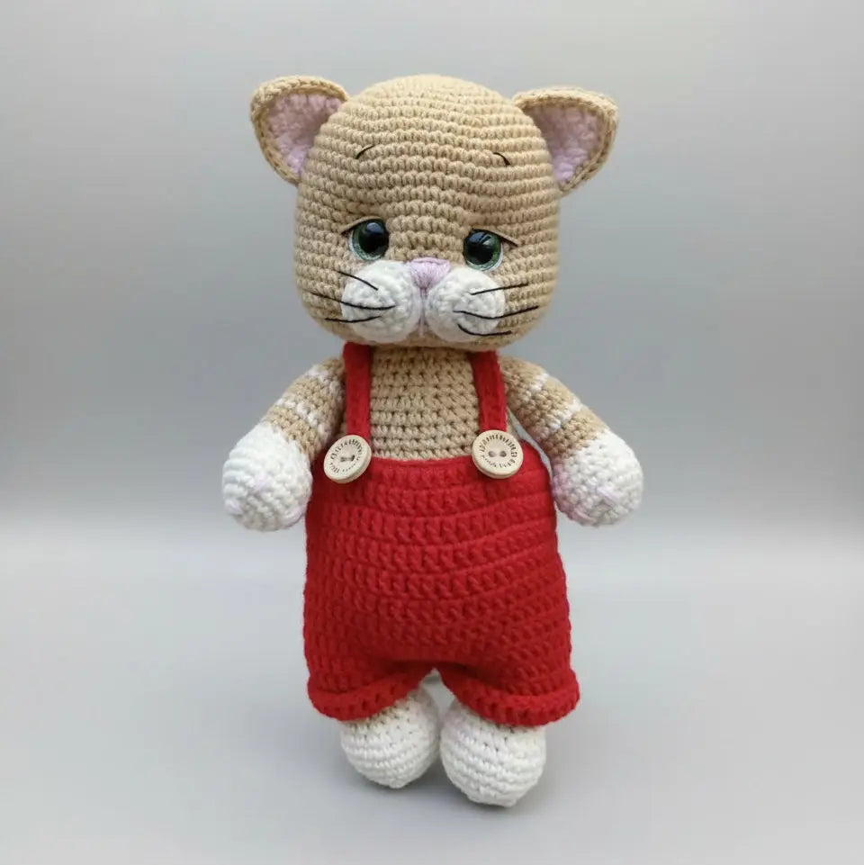 Crocheted cat plushie in red overalls, standing on a neutral background