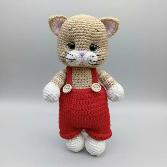 Crocheted cat plushie in red overalls, standing on a neutral background