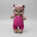 Crocheted teddy bear in pink overalls with flower crown, standing on gray background